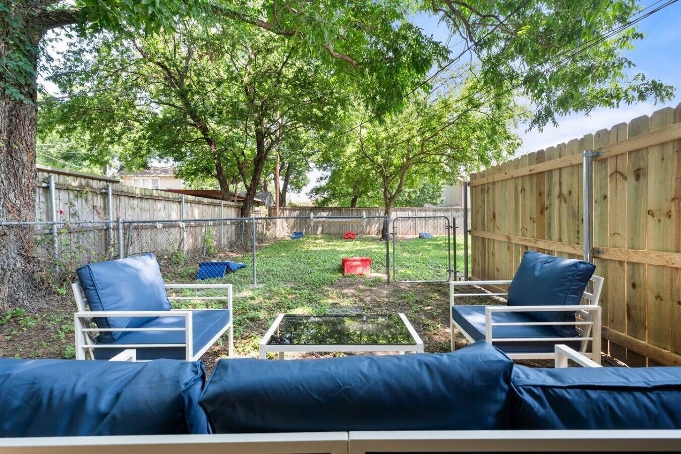 Un espace extérieur aménagé avec deux chaises en métal blanc et des coussins bleus, entouré d’une clôture en bois et d’arbres.