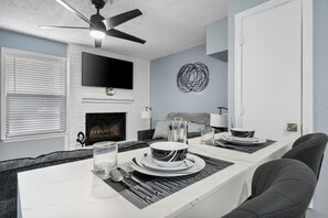 A modern dining area with a white table set for four, a fireplace, and a ceiling fan. - Game Day Modern 2BR | Steps to AT&T & Globe Life (Arlington)