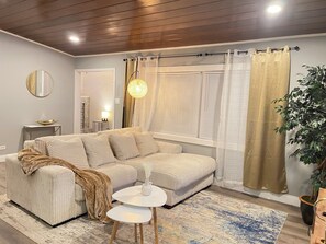 Ein modernes Wohnzimmer mit einem Ecksofa, einem kleinen runden Tisch, einer Topfpflanze und einem großen Fenster mit Vorhängen.