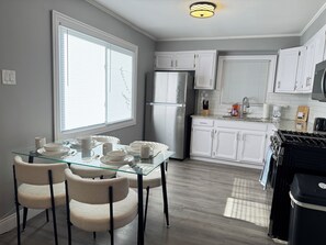 Une cuisine moderne avec des meubles blancs, un réfrigérateur en acier inoxydable, une cuisinière à gaz et un coin repas comprenant une table et des chaises en verre.