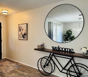 Un couloir avec un miroir rond, un panneau d’accueil, un vélo et une petite plante.