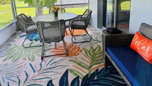A covered patio with a round table and four chairs, a bench with a cushion, and a view of a house and lawn. - Stylish Lakefront Retreat, Pet Friendly, Flexible King/Twin Setup, Mins to Beach (Venice)