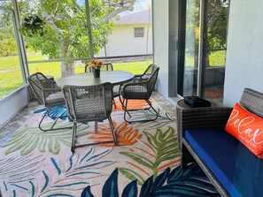 Eine überdachte Terrasse mit einem runden Tisch, vier Stühlen und einer Sitzbank mit Kissen, mit Blick auf ein Haus und eine Rasenfläche.