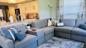 A modern living room with a sectional sofa, a kitchen area, and a large window with curtains. - Stylish Lakefront Retreat, Pet Friendly, Flexible King/Twin Setup, Mins to Beach (Venice)