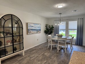 A dining area with a round table, white chairs, a large plant, and a painting of a landscape. - Stylish Lakefront Retreat, Pet Friendly, Flexible King/Twin Setup, Mins to Beach (Venice)