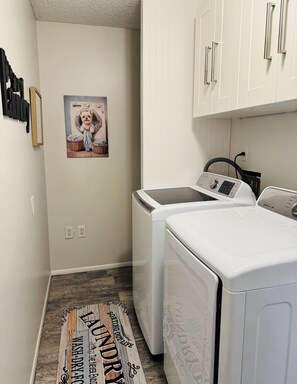 A laundry room with two front-loading washers and dryers, a wall-mounted drying rack, and a poster of a dog in a laundry setting. - Stylish Lakefront Retreat, Pet Friendly, Flexible King/Twin Setup, Mins to Beach (Venice)