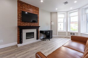 A modern living room with a brick fireplace, a flat-screen TV mounted above it, a black office chair, a brown leather sofa, and large windows allowing natural light. - Bright & Spacious Apartment in Brooklyn (Brooklyn)
