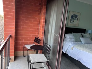 A balcony with a brick wall, two chairs, a table, and a bed with pillows and a blanket. - A comfortable apartment with a charming view over Adelaide’s southern parklands. (Adelaide)