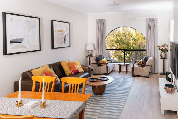 Ein modernes Wohnzimmer mit einer Couch, Sesseln, einem Couchtisch und einem Esstisch. Zwei gerahmte Kunstwerke hängen an der Wand, ein Fenster mit Vorhängen und ein Blick auf Bäume im Freien.