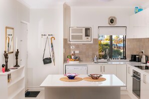 A modern kitchen with white cabinetry, a built-in dishwasher, a window with a view, and a small island with a bowl and a bottle. - A comfortable apartment with a charming view over Adelaide’s southern parklands. (Adelaide)