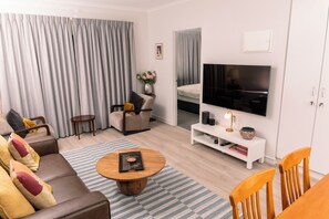 A modern living room with a sofa, coffee table, TV, and a dining area with chairs. - A comfortable apartment with a charming view over Adelaide’s southern parklands. (Adelaide)
