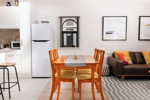 A modern kitchen with white cabinets, a white refrigerator, and a microwave. There is a dining table with chairs and a window above it. A brown sofa and a rug are in the living area. - A comfortable apartment with a charming view over Adelaide’s southern parklands. (Adelaide)