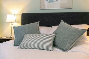 A neatly made bed with a dark headboard, multiple pillows, and a bedside lamp. - A comfortable apartment with a charming view over Adelaide’s southern parklands. (Adelaide)
