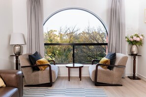 A room with two armchairs, a small table, a lamp, and a view of trees through a large window. - A comfortable apartment with a charming view over Adelaide’s southern parklands. (Adelaide)