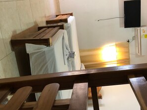 A room with a bed, a wooden bench, a television mounted on the wall, and a window with sunlight.