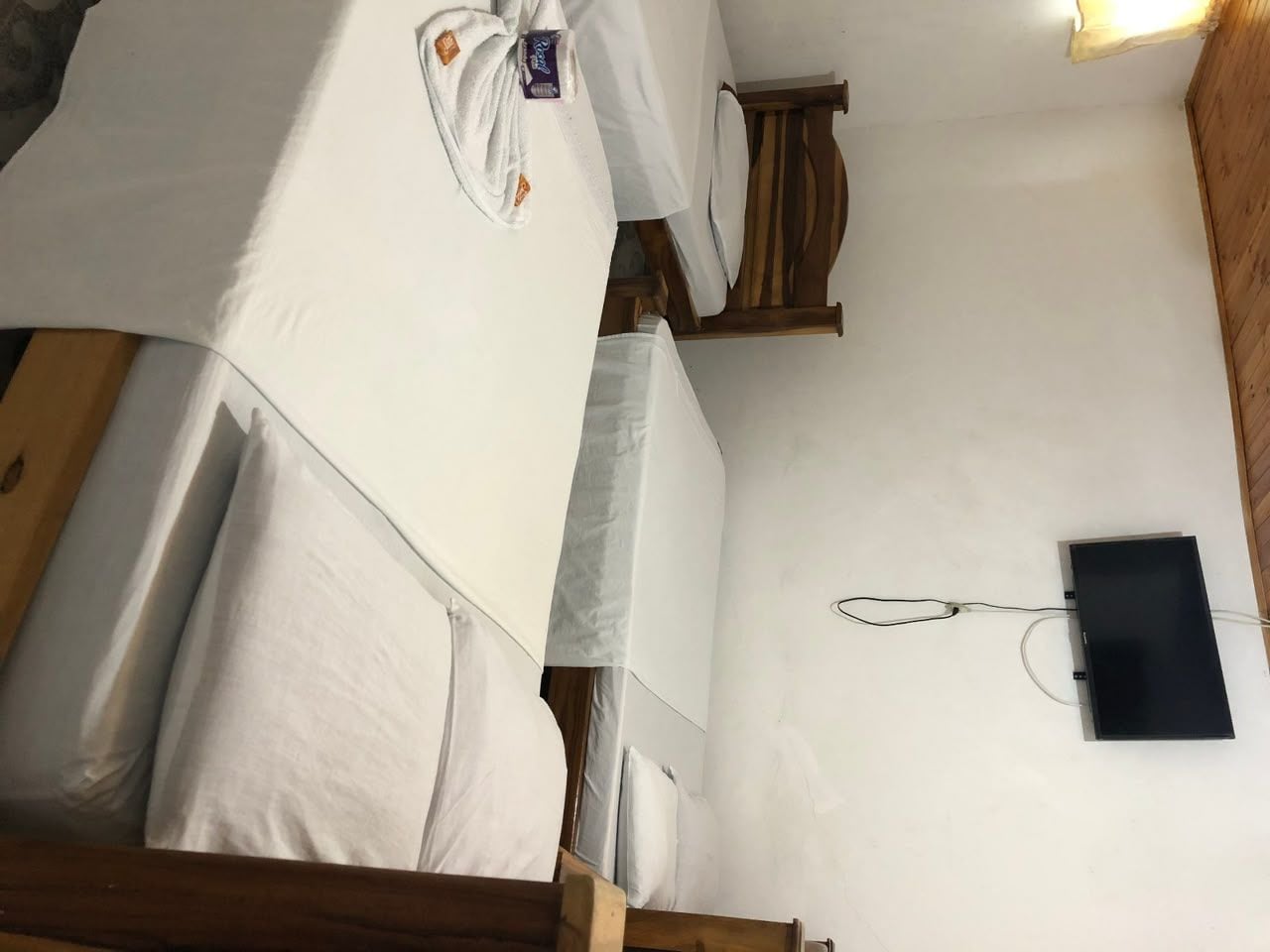 Una habitación con dos camas, un cabecero de madera, un objeto negro pequeño en la pared y una pared blanca.