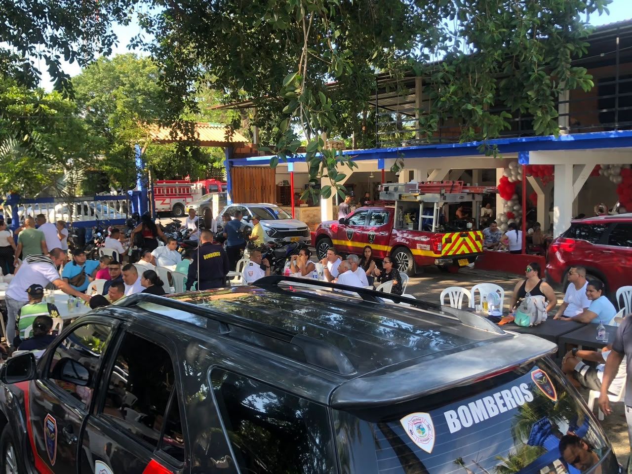 Un evento al aire libre concurrido, con gente sentada en mesas, un vehículo negro con la leyenda 'BOMBEROS' y un vehículo rojo al fondo.