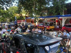 A crowded outdoor event with people seated at tables, a black vehicle with 'BOMBEROS' text, and a red vehicle in the background.