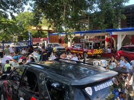 Un evento al aire libre concurrido, con gente sentada en mesas, un vehículo negro con la leyenda 'BOMBEROS' y un vehículo rojo al fondo.