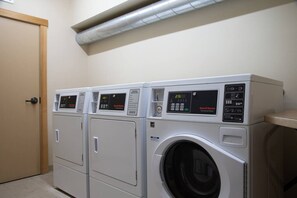 A laundry facility with three stacked washers and dryers, control panels, and a ventilation duct above. - Ski-In King Executive Resort Suite w/ Mtn Views, Pool & Garage! (Mammoth Lakes)