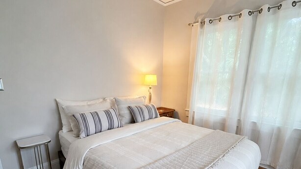 A bedroom with a bed, bedside table, lamp, and a view of the outdoors through sheer curtains. - The Natty - Charming 3BR Bungalow Near UF, The Swamp & Law School (Gainesville)