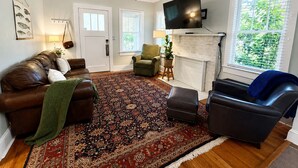 A living room with a brown leather sofa, a green armchair, a black leather chair, a patterned rug, a ceiling fan, a flat-screen TV, and a fireplace. - The Natty - Charming 3BR Bungalow Near UF, The Swamp & Law School (Gainesville)