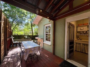 Eine Holzterrasse mit Tisch und Stühlen, ein überdachter Außenbereich und eine durch eine offene Tür sichtbare Küche.