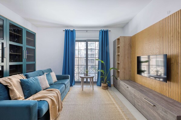 A living room with a sofa, TV, wooden panel wall, and a view through a window. - Belmonte Heights Flat 2BDR (Sliema)