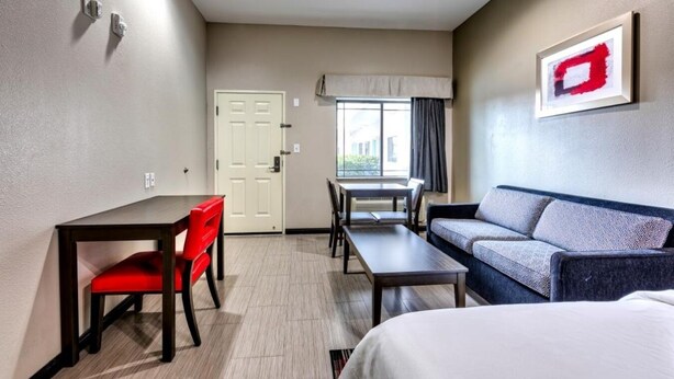 A hotel room with a bed, a sofa, a desk, and a chair. There is a window with curtains, a door, and a wall-mounted artwork.