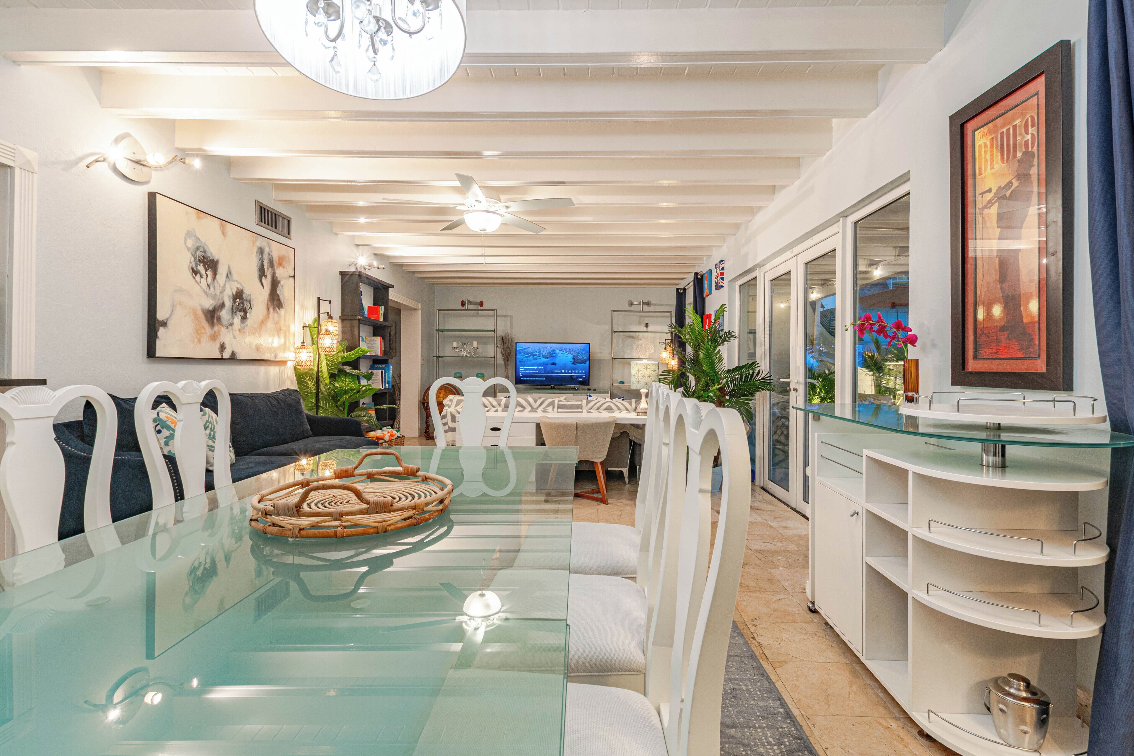 A modern living room with a glass dining table, white chairs, a ceiling fan, and a television.
