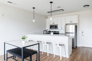 Een moderne keuken met witte kastjes, een centraal eiland met barkrukken, een zwarte eettafel en een potplant.