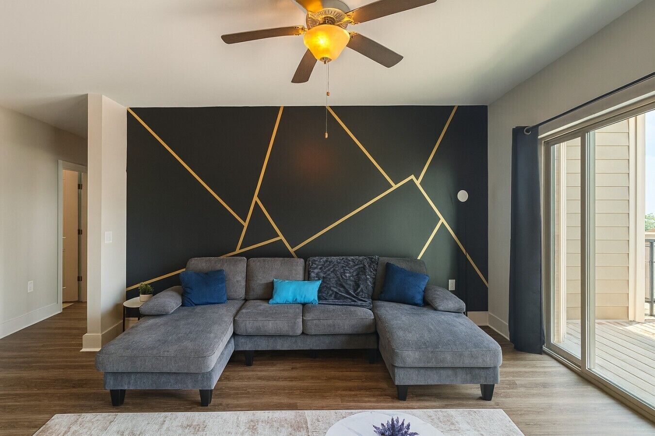 A modern living room with a sectional sofa, a ceiling fan, and a geometric wall design.