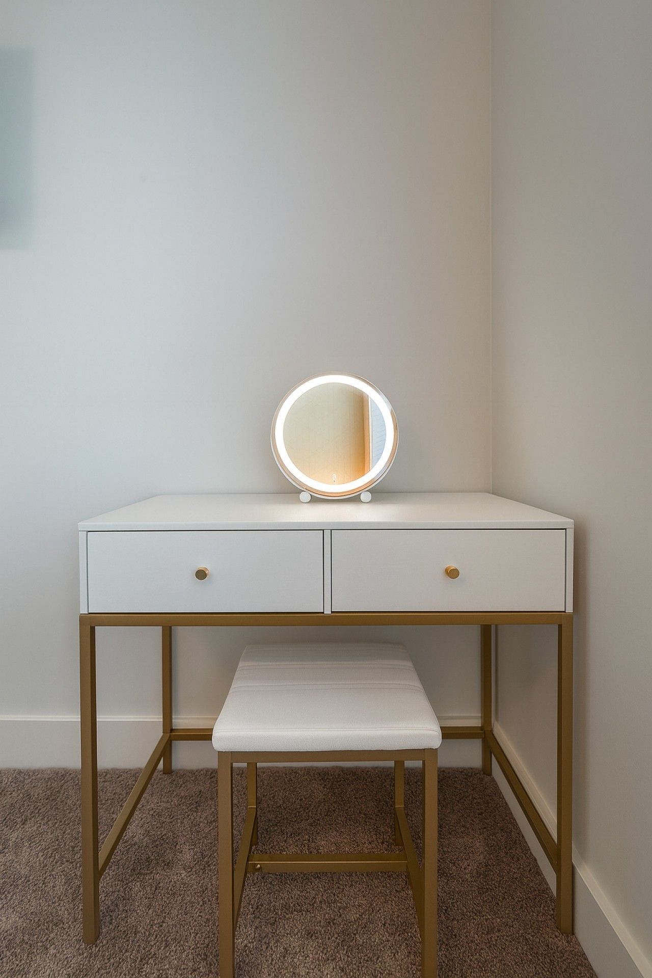 A white vanity with a round mirror and two drawers, a bench, and a gold frame.