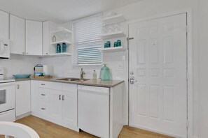 Una cocina compacta con armarios blancos, encimera de granito, lavavajillas integrado y una ventana con persianas.