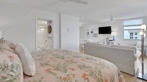 Un dormitorio con cama, ventilador de techo, baño con inodoro y kitchenette con televisor.