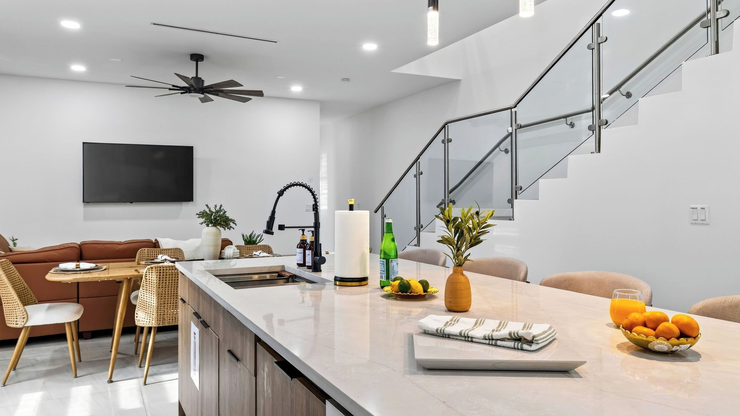 Una cocina moderna con una isla central, un área de comedor con sofá y sillas, y una escalera con barandillas de vidrio.