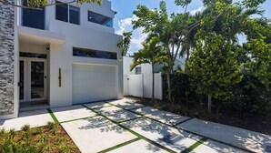 Una moderna casa a due piani con garage, facciata in pietra e un giardino curato con palme.