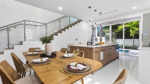 A modern kitchen with a wooden dining table set for four, a built-in island with a sink, and a staircase with glass railings. - Park & Palm by Avantstay Private Pool + 4 Ensuite Baths + BBQ Victoria Park + Las Olas (Fort Lauderdale)