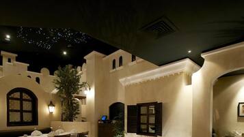 An outdoor dining area with tables set for two, surrounded by walls with decorative elements and a ceiling with a starry light display.