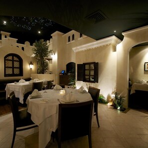 Un área de comedor al aire libre con mesas dispuestas para dos, rodeada de muros con detalles decorativos y un techo con una iluminación que simula estrellas.