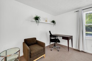 A room with a brown armchair, a desk with a chair, a glass table, and two hanging plants on a white shelf. - Spacious 4BR Home •Near DC  •Free Parking and Fast Wi-Fi (Silver Spring)