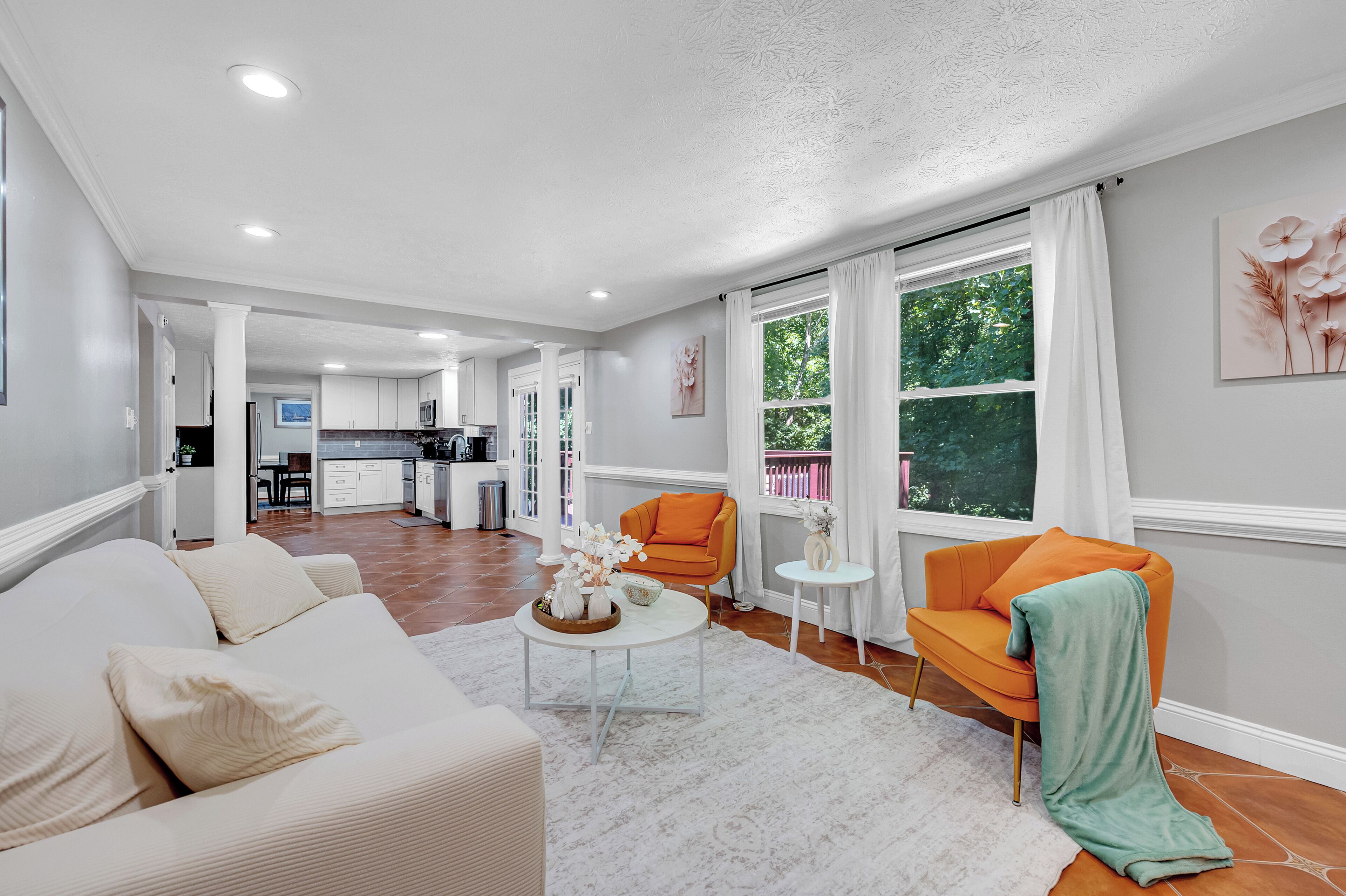 A modern living room with a sofa, armchairs, a coffee table, and a view of the kitchen and outdoor area.