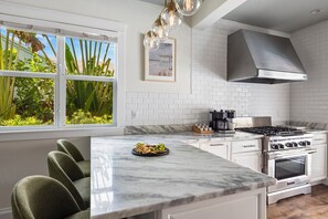 A modern kitchen with white subway tiles, a marble countertop, a stainless steel stove, and a large window with a view of greenery. - Lux WPB Oasis with Heated Pool/Spa + GameRoom – Sleeps 12! (West Palm Beach)