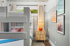 A bunk bed room with a wooden nightstand and a lamp, framed artwork on the wall, and a window with curtains. - Lux WPB Oasis with Heated Pool/Spa + GameRoom – Sleeps 12! (West Palm Beach)