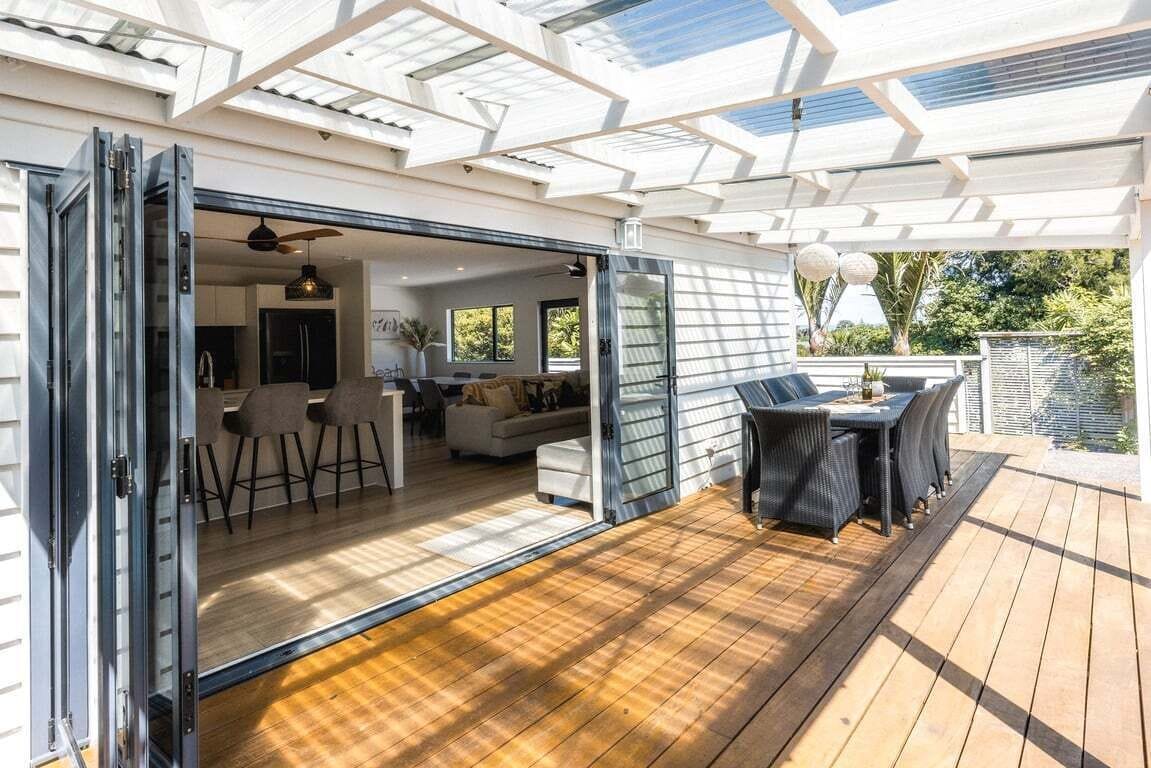 Una casa moderna amb zona de pati que inclou una taula i caires de menjador, i una porta corredissa de vidre que dona a un espai interior amb cuina i zona de seients.