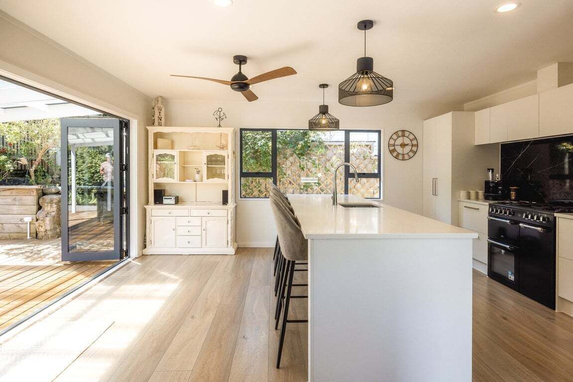 A modern kitchen with a central island, bar stools, a ceiling fan, pendant lights, and a view through a glass door to an outdoor area.