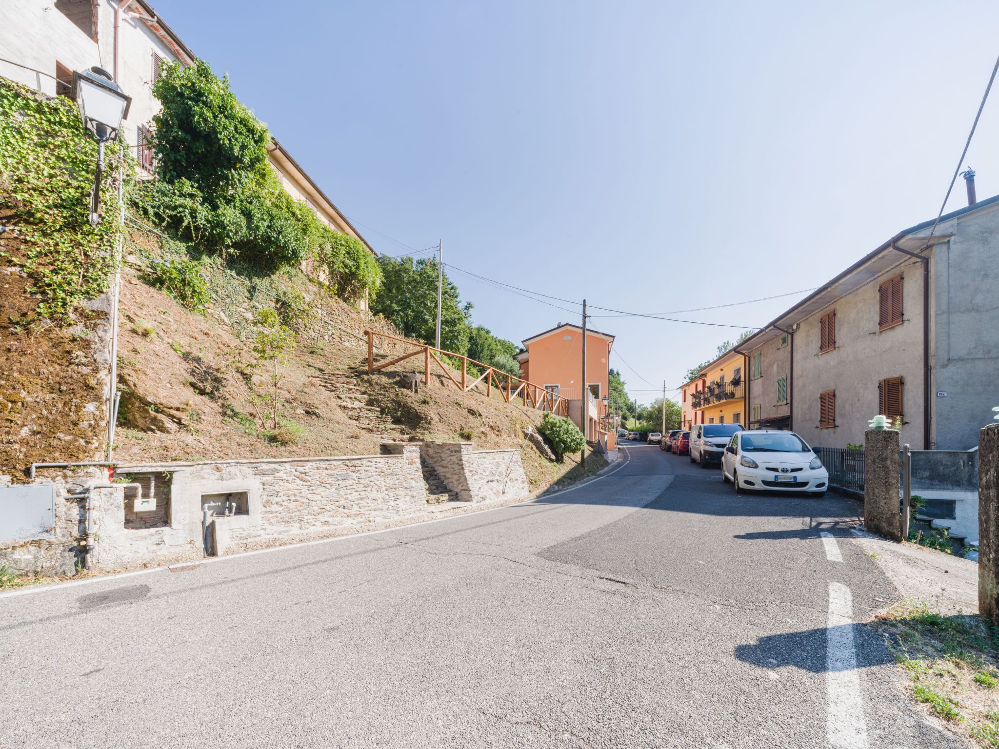 Una stretta via fiancheggiata da edifici, un muro in pietra alla sinistra e auto parcheggiate a destra.