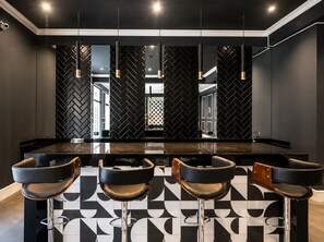 Un área de bar moderna con piso de baldosas geométricas en blanco y negro, una barra central y cuatro taburetes de bar.
