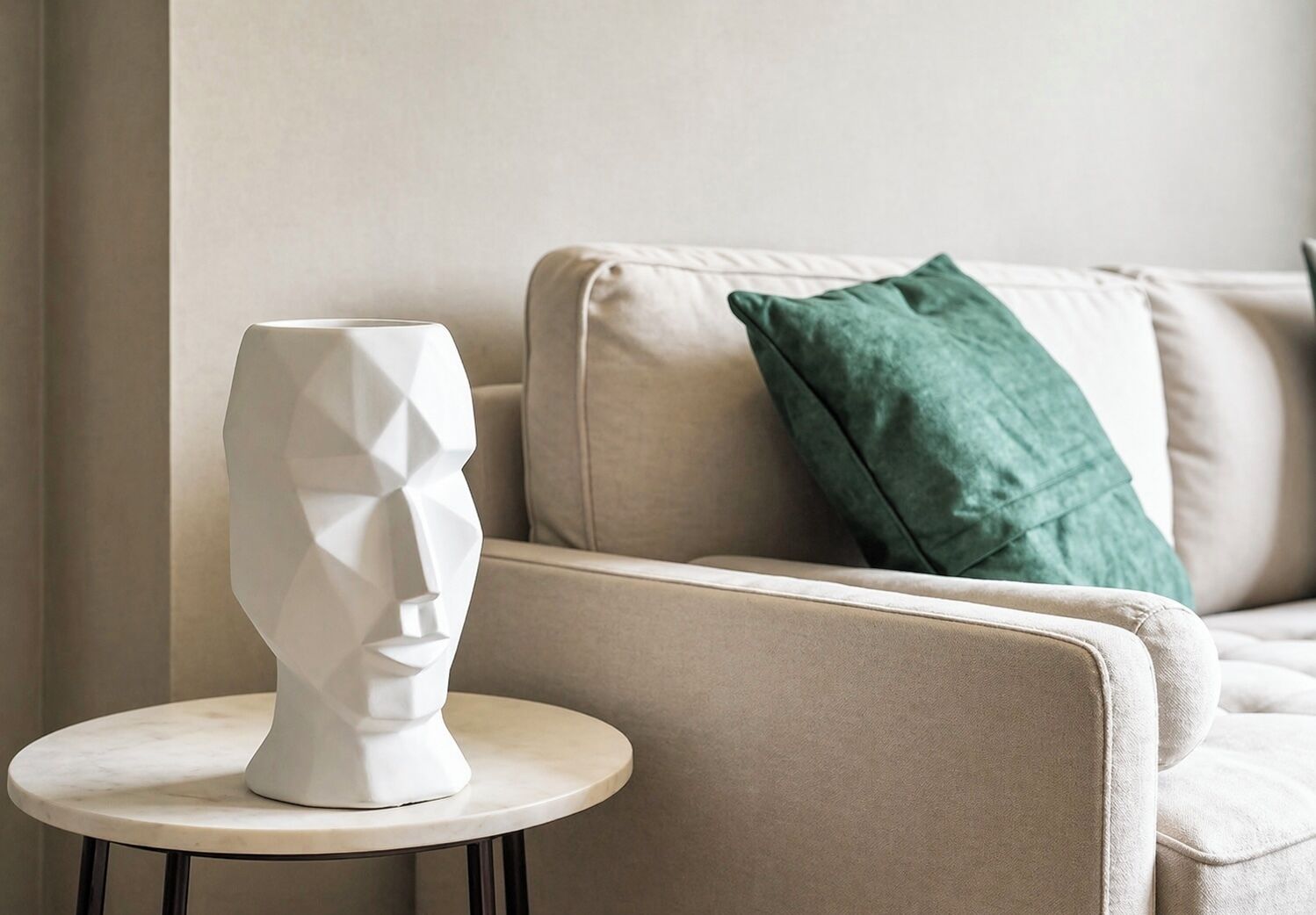 A white geometric sculpture on a round table beside a beige sofa with a green pillow.