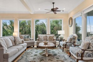 A spacious living room with a ceiling fan, large windows with a view of palm trees, and a mix of sofas and chairs. - Gypsy Tide! Luxury 5 BR, 5.5 BA Oceanfront Home-Folly Beach! (Folly Beach)